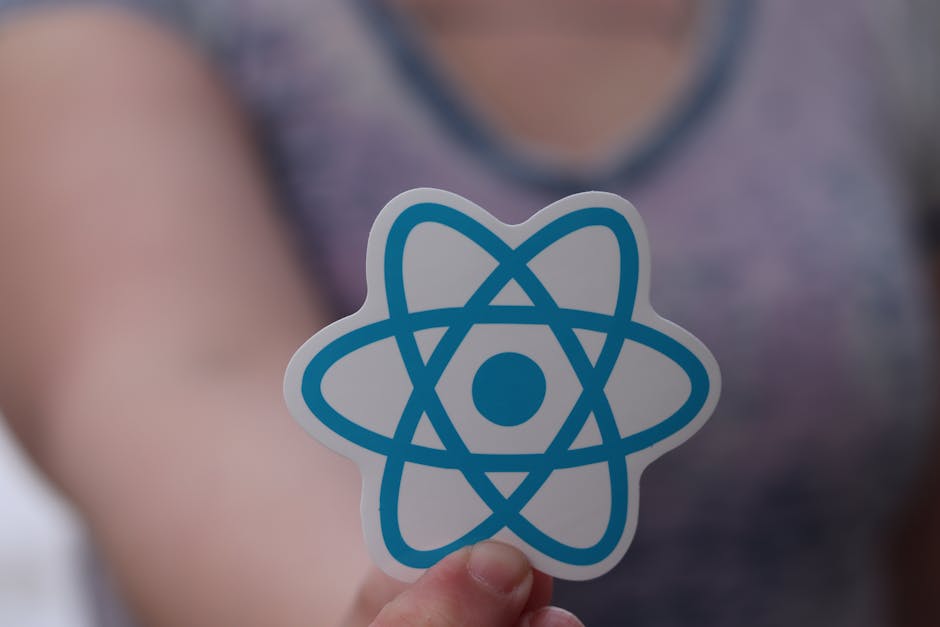 A person holding a sticker featuring a blue atomic symbol with a central circle and interconnected loops, set in focus in the foreground. In the background, part of a person dressed in a casual top is