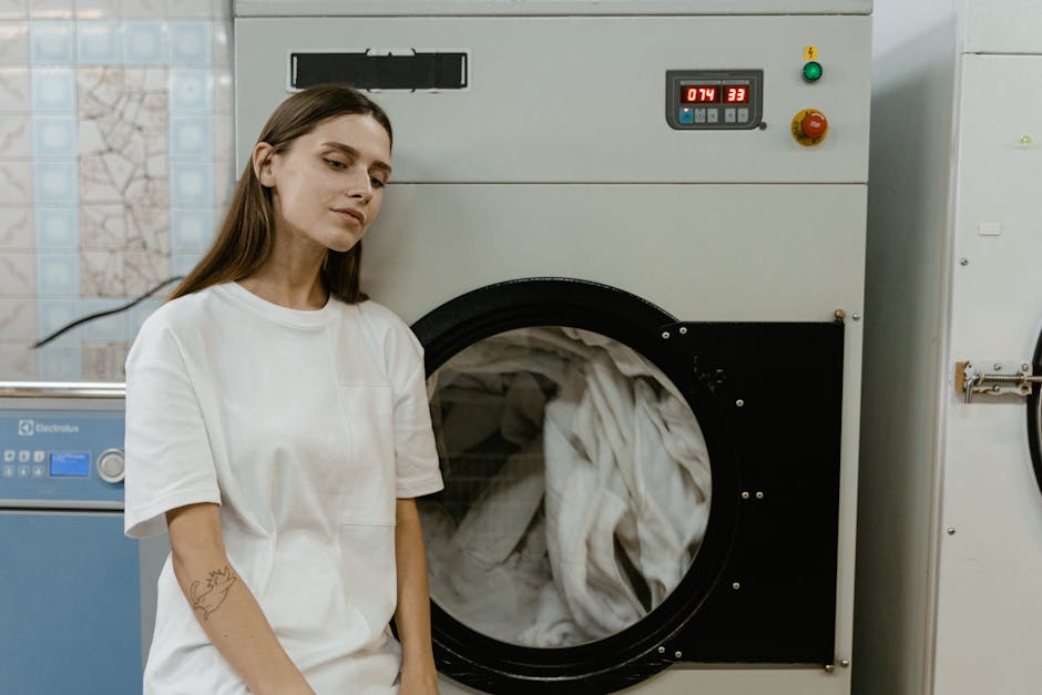A young woman dressed in a white uniform is standing in a commercial laundry room, next to a large industrial washing machine filled with white linen. The machine has a black circular door through whi